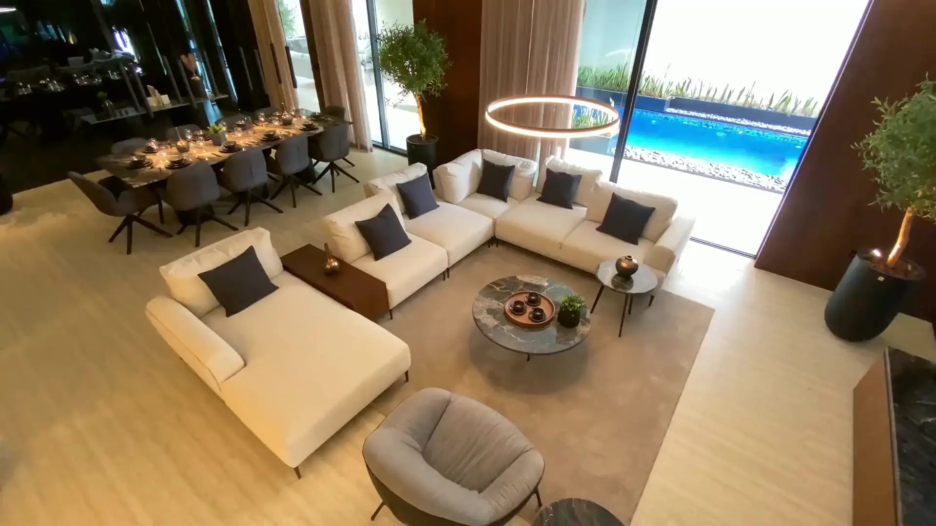 Sunlit double-height living room with ivory sofas, a ring pendant light and a pool beyond the glass doors