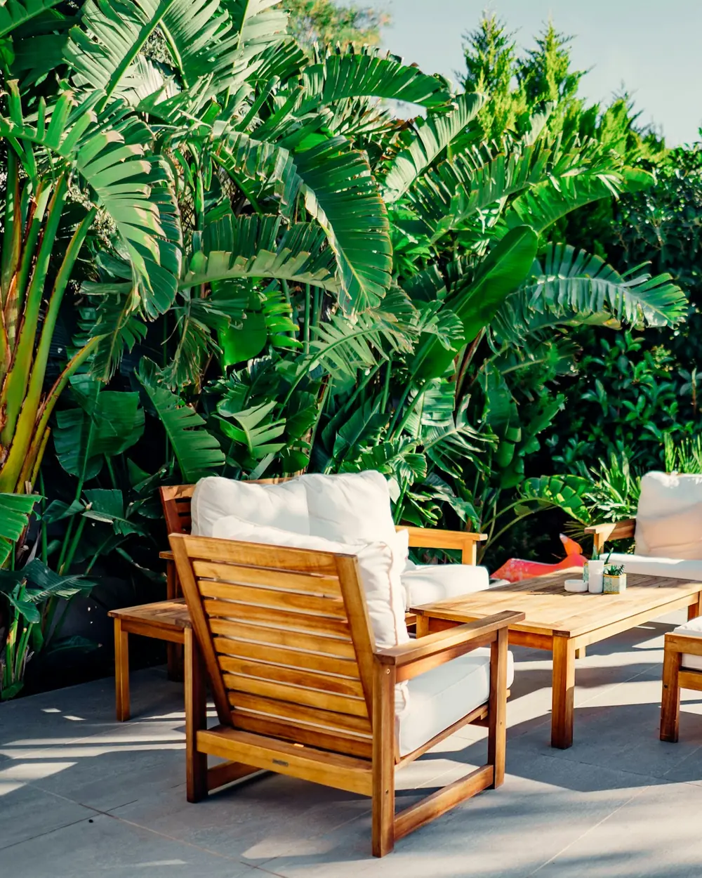 Teak outdoor lounge chairs with ivory cushions on a terrace framed by tropical plants
