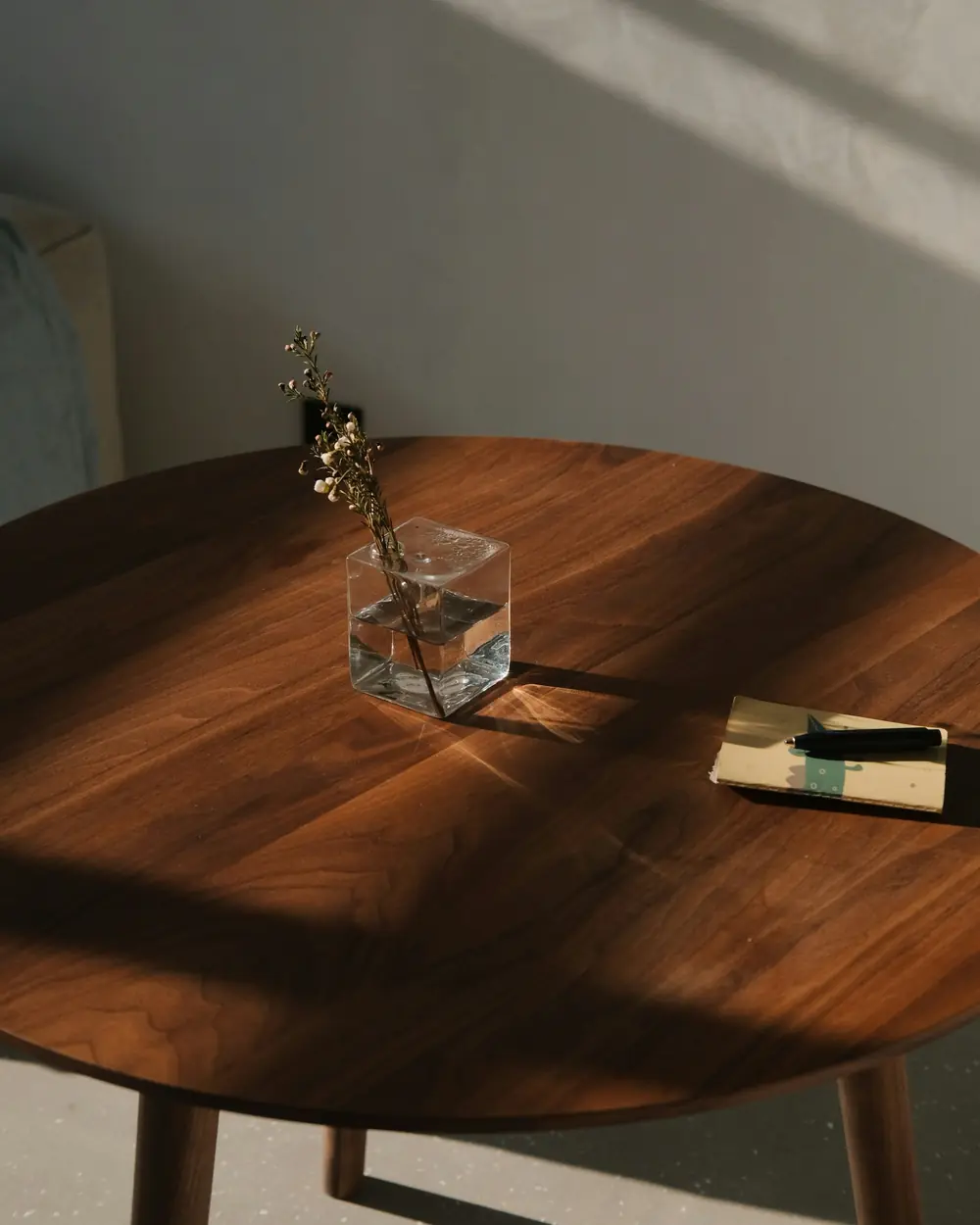 Round walnut-toned coffee table in raking afternoon light with a glass vase and a book