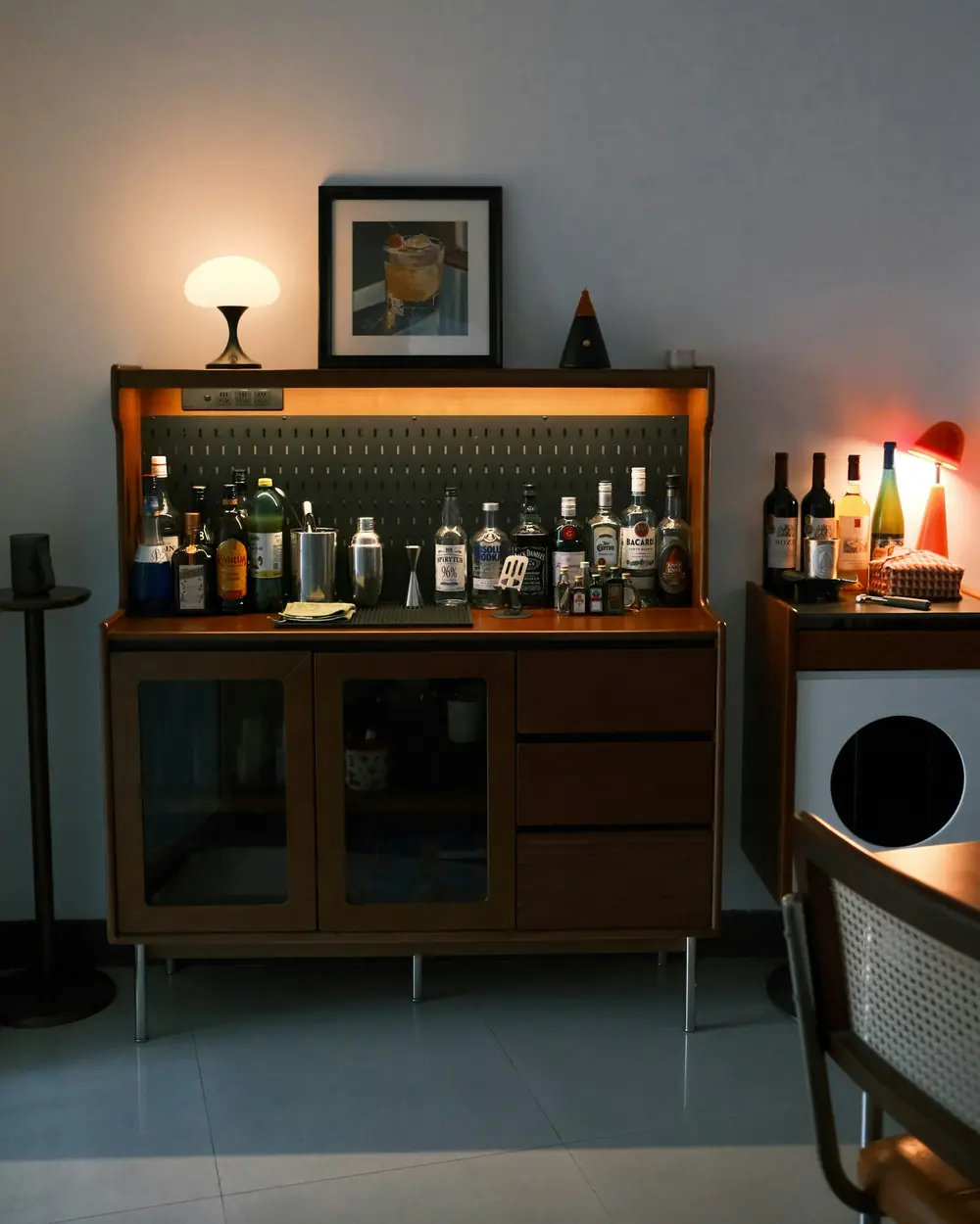 Walnut bar cabinet with glass doors and a lit bottle shelf, warm table lamp on top
