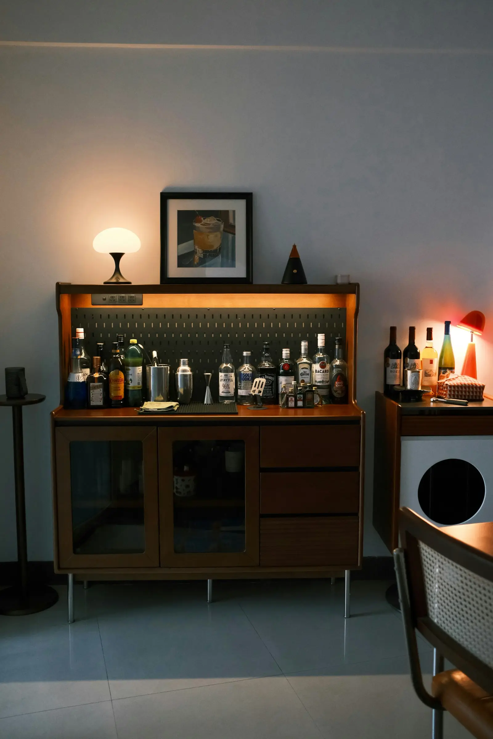 Walnut bar cabinet with glass doors and a lit bottle shelf, warm table lamp on top