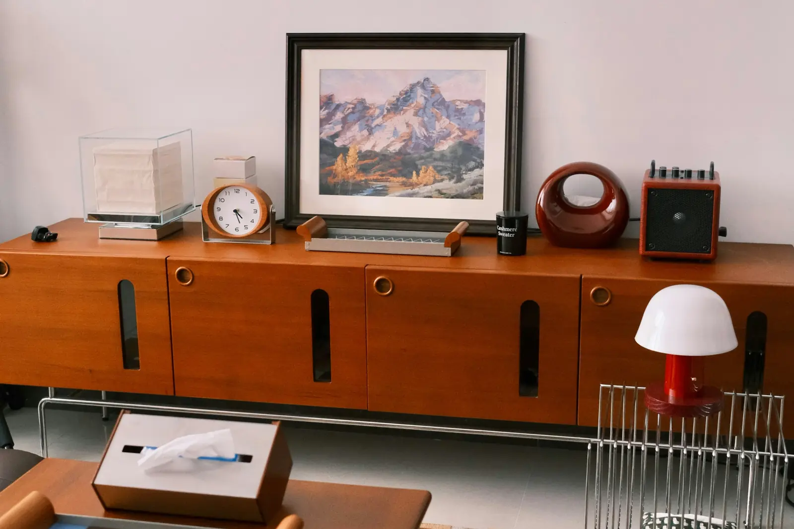 Teak sideboard with a framed mountain print, a table clock and a red mushroom lamp